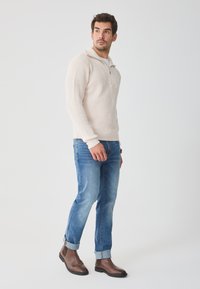 Világos bézs bordázott pulóver magas gallérral, közepes mosású kék farmernállal és barna bőr bokacsizmával párosítva. Tiszta és minimalista dizájn.