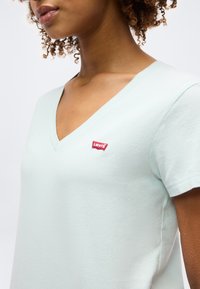 Γυναίκα που φοράει ανοιχτό μπλε μπλουζάκι V-neck με μικρό κόκκινο λογότυπο Levi's στο στήθος, κομμένο από τους ώμους έως τη μέση του κορμού.