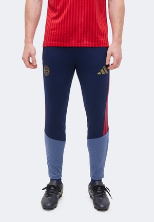 Mand iført marineblå og lyseblå træningsbukser med røde striber, sorte fodboldstøvler og en rød sports t-shirt.