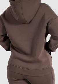 Osoba w jednolitym brązowym hoodie i pasujących spodniach, stojąca tyłem z dłońmi w kieszeniach, na neutralnym tle.