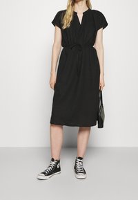 Robe noire, arrivant au genou, avec des manches courtes et un col en V. Présente un cordon ajustable à la taille. Assortie avec des baskets montantes.