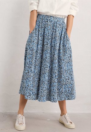 Femme portant une jupe midi à fleurs bleues et blanches avec des poches et des baskets blanches, debout contre un mur clair uni.