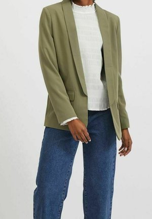 Person iført en olivengrøn blazer over en hvid tekstureret bluse og blå denimjeans mod en ensfarvet baggrund.