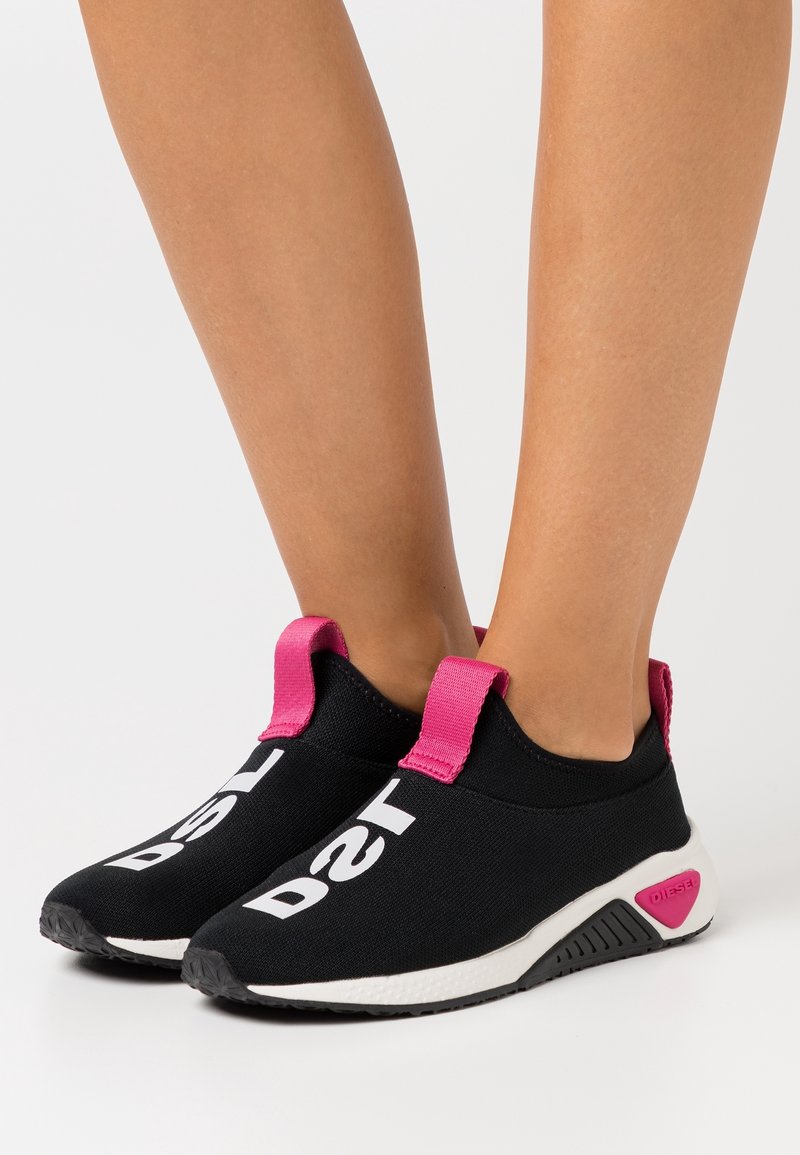 Baskets slip-on noires à semelles blanches, avec un grand texte blanc sur le dessus et des languettes de tirage rose vif au talon.