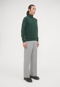 Donkergroene gebreide pullover met een hoge kraag en rits, gedragen met lichtgrijze wijde broek en zwarte stevige schoenen. Eenvoudig ontwerp, geen patronen.