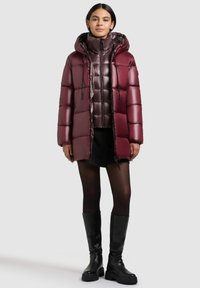 Burgundy pufferjacka med huva och dragkedja, med en glänsande yta och quiltat mönster. Buren över en svart outfit och knähöga stövlar.