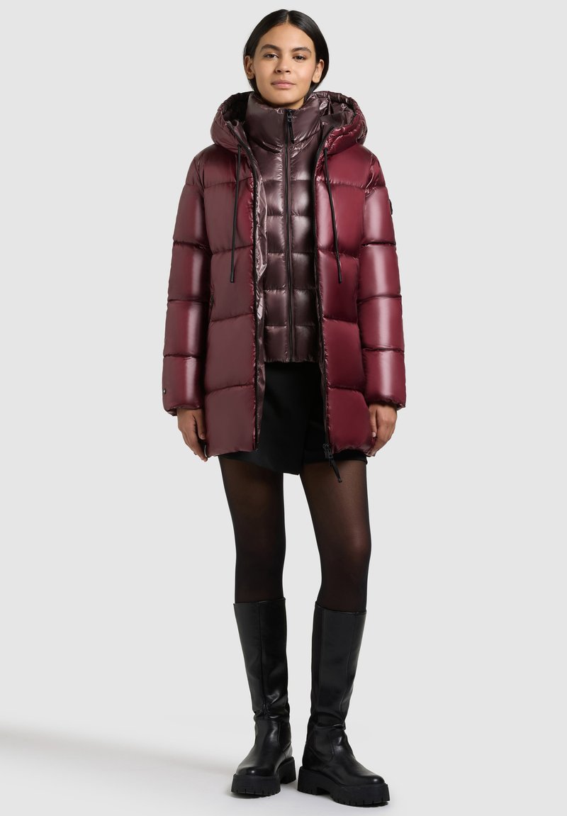 Burgundy pufferjacka med huva och dragkedja, med en glänsande yta och quiltat mönster. Buren över en svart outfit och knähöga stövlar.