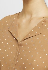 Camicetta marrone a pois con colletto, caratterizzata da piccoli pois bianchi distribuiti in modo uniforme. Tessuto liscio, chiusura a bottoni al collo.