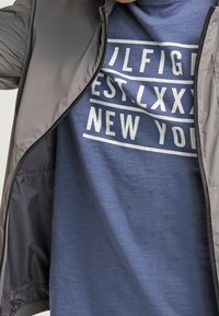 Persona vestida con una camiseta azul con texto blanco, parcialmente cubierta por una chaqueta gris con cremallera, enfocándose en el texto de la camiseta y la parte superior del torso.