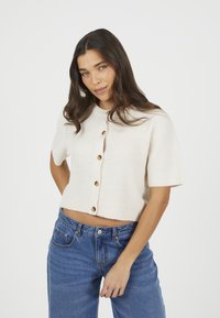 Jasno beżowy, krótki sweter współczesny z dzianiny z prążkowaną teksturą i pięcioma brązowymi guzikami. Noszony z dżinsami w kolorze niebieskim i wysokim stanem.