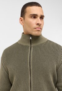 Olivgrön zip-up kofta med ribbad textur, hög krage och kontrasterande mörka ränder. Har en slät dragkedja och lättviktig tyg.