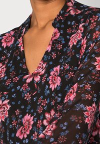 Gros plan sur une personne portant un blouse noire avec des motifs floraux roses et bleus et un décolleté en V.