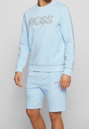 Mann trägt ein hellblaues Sweatshirt mit dem Schriftzug "BOSS" und passende Shorts mit kleinem "BOSS" Logo am linken Oberschenkel.