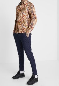 Chemise à motifs multicolores avec un col, associée à un pantalon navy à rayures et des baskets noires. Texture de tissu lisse.