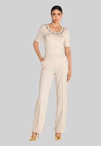 Vrouw in een beige top met korte mouwen en bloemmotief bij de hals, bijpassende hoge taille broek, gouden hoge hakken, staand met één hand in haar zak.
