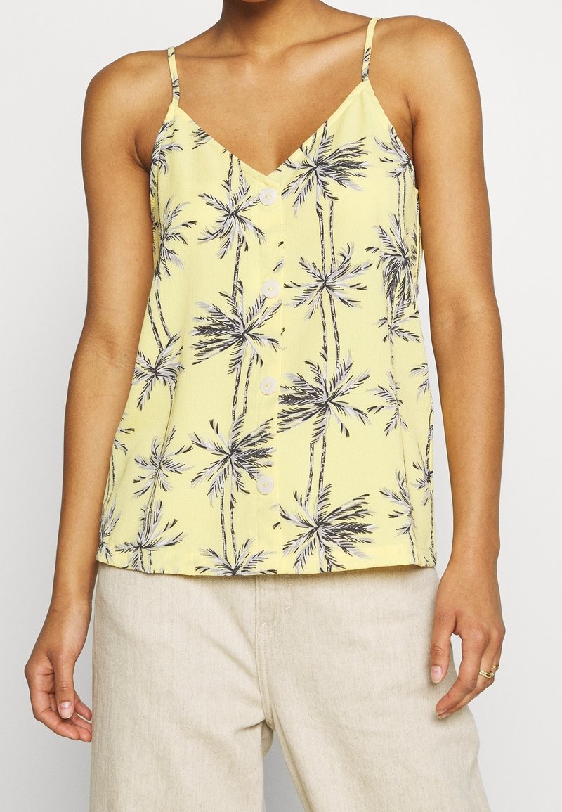 Haut jaune sans manches boutonné avec imprimé de palmiers noirs porté avec un pantalon beige sur une personne aux bras détendus.