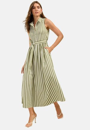 Frau trägt ärmelloses, knöchellanges Kleid mit Kragen und vertikalen grünen und weißen Streifen, steht mit einer Hand in der Tasche und trägt Absätze.