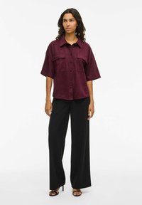 Bordo krāsas oversize krekls ar īsām rokām, divām priekšējām kabatām un pogu aizdari, kopā ar melnām platajiem biksēm un augstpapēžu sandālēm.