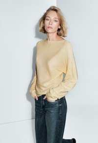 Giovane donna con capelli biondi corti, che indossa un maglione giallo chiaro e jeans blu scuro, in piedi con le mani nelle tasche su uno sfondo bianco.