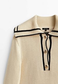 Pull en tricot couleur crème avec un large col bordé de noir, fermeture à trois boutons et un motif de tissu texturé.