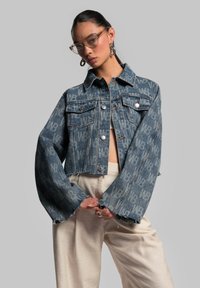 Gecutte Jeansjacke in einem verblassten blauen Farbton, mit einem Logomuster, zwei Brusttaschen und ausgefransten Ärmeln. Trägt sie über hellen Hosen.