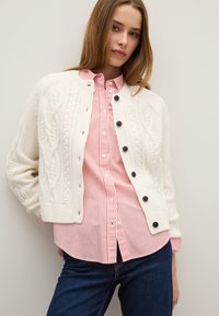 Cremfarbener Zopfmuster-Cardigan mit schwarzen Knöpfen über einem rosa-weißen gestreiften Hemd, kombiniert mit dunklen blauen Jeans. Gerade Passform.