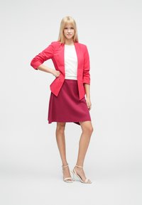 Blazer rosso con maniche a tre quarti sopra una maglietta bianca, abbinato a una gonna a trapezio bordeaux. La modella indossa tacchi nude con cinturini.