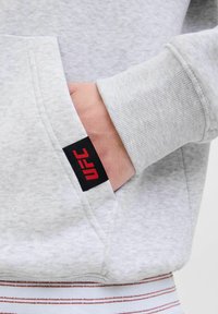 Hand in der Tasche eines hellgrauen Sweatshirts mit UFC-Logo, das einen gerippten Bund hat, mit einem gestreiften Hemd, das darunter sichtbar ist.