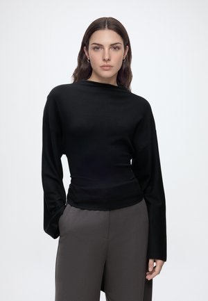 Jeune femme portant un haut noir cintré à manches longues et un pantalon gris ample, debout devant un fond blanc uni.