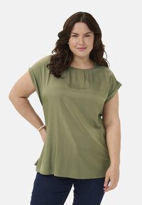 Bluză scurtă, verde măsliniu, cu guler rotund și siluetă fluidă; textură netedă și cădere moale, asortată cu blugi de culoare albastru închis.