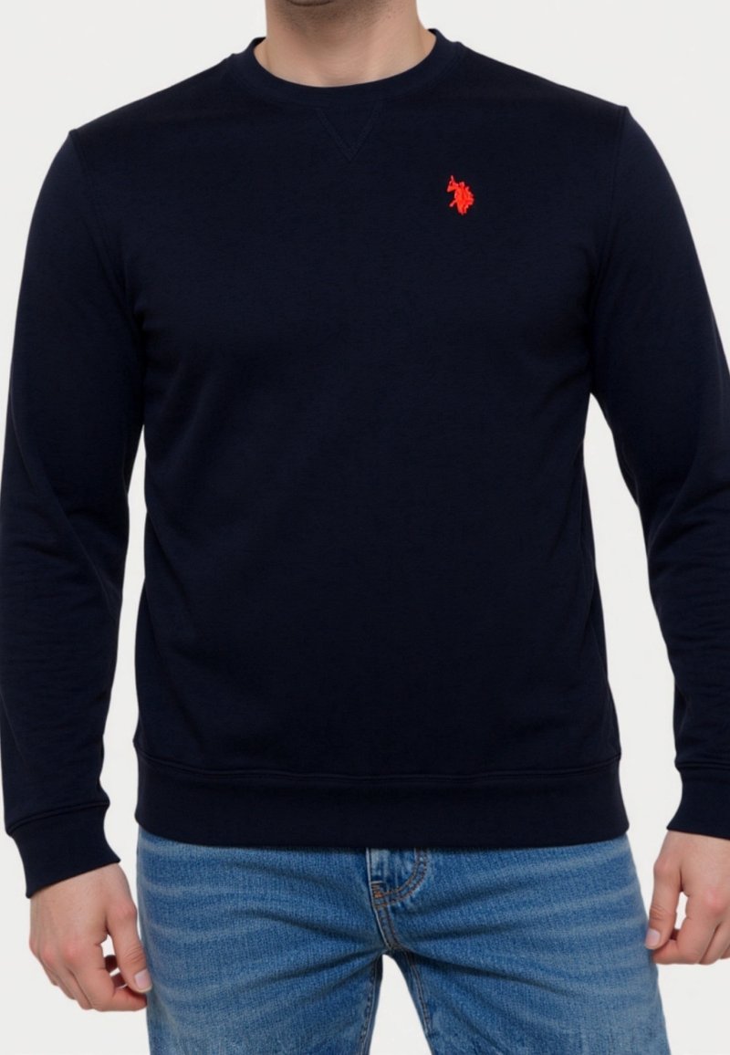 Mann trägt ein eng anliegendes schwarzes Sweatshirt mit langen Ärmeln und einem roten, auf der linken Brust eingestickten Logo sowie blaue Jeans aus Denim.
