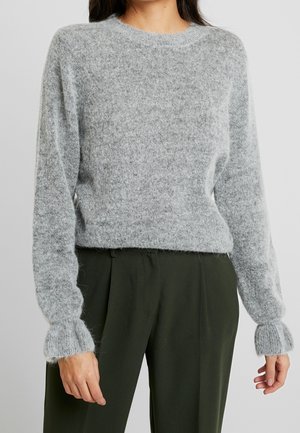 Femme portant un pull en maille duveteuse gris clair avec des poignets côtelés et un pantalon taille haute vert olive foncé.