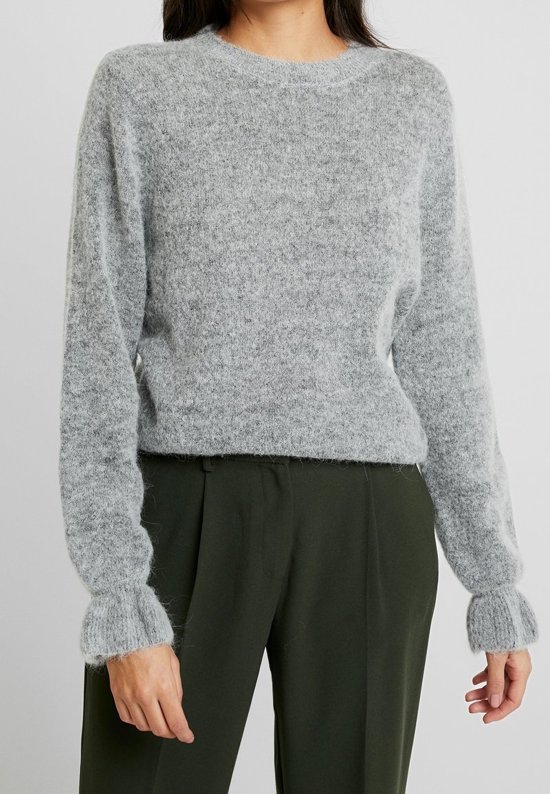 Femme portant un pull en maille duveteuse gris clair avec des poignets côtelés et un pantalon taille haute vert olive foncé.