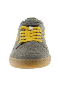 Baskets en suède gris avec des lacets et des accents jaunes, présentant un design perforé sur le bout, et une semelle en caoutchouc marron.