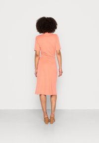 Žena s kudrnatými vlasy, oblečená v korálových midi šatech a světle hnědých blokových podpatcích, stojí čelem k jednobarevnému světle šedému pozadí.