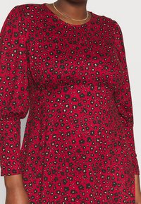 Robe rouge avec un motif léopard en noir et beige, manches longues bouffantes et encolure ronde, confectionnée en tissu doux et lisse.