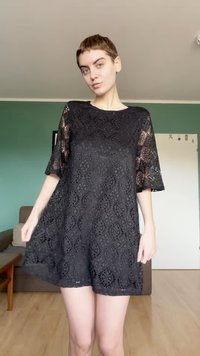 Jeune personne aux cheveux courts portant une robe en dentelle noire, debout dans une pièce aux murs verts et au sol en bois.
