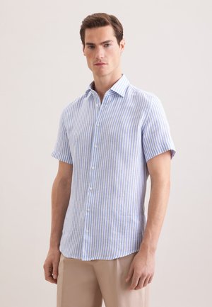 Homme portant une chemise à boutons à manches courtes à rayures verticales bleu clair et blanc avec un pantalon beige, debout devant un fond uni.
