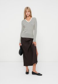 Gestreepte langemouwen top in crème en zwart, gecombineerd met een donkerbruin midi-rok met een zijsplit, en zwarte flats met een ronde neus.