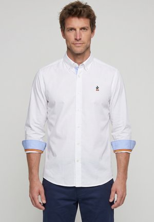 Hombre con camisa blanca de botones con puños azul claro enrollados y pantalones azul marino, de pie frente a un fondo liso de color gris claro.