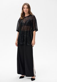 Kobieta w przezroczystej czarnej bluzce z cekinami nosi czarny stanik, szerokie czarne spodnie z białymi bocznymi paskami oraz czarne czółenka na ostrym nosku, na białym tle.