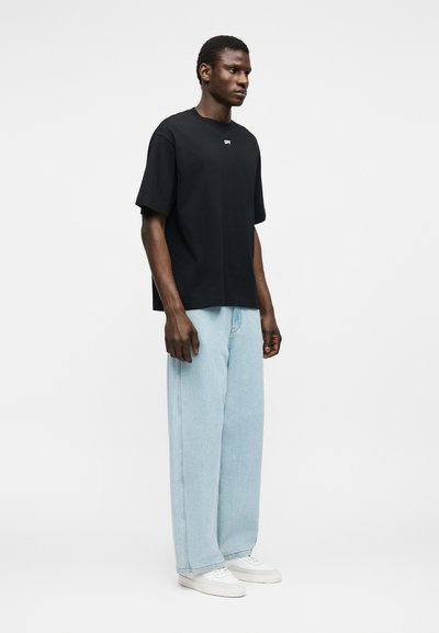 Άνδρας που φορά μαύρο φαρδύ T-shirt με μικρό λευκό κείμενο, ανοιχτό μπλε τζιν παντελόνι φαρδιάς γραμμής και λευκά αθλητικά παπούτσια, στέκεται μπροστά σε απλό φόντο.