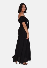 Abito maxiuniforme nero con spalle scoperte, maniche a palloncino corte, corpetto aderente e gonna fluida. Presenta un dettaglio con fiocco in vita.