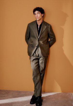 Joven con traje verde oliva y zapatos negros apoyado contra una pared color beige con las manos detrás de la espalda, proyectando una sombra.