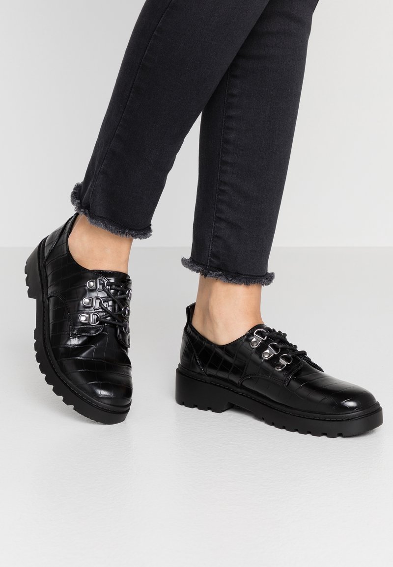 Chaussures noires en cuir embossé crocodile à lacets, portées avec un pantalon noir court à ourlets effilochés sur un sol gris clair.