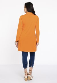 Langærmet orange tunika med krave-design, der har sideskår. Sammen med denim leggings og brune kilehæle med ankelremme.