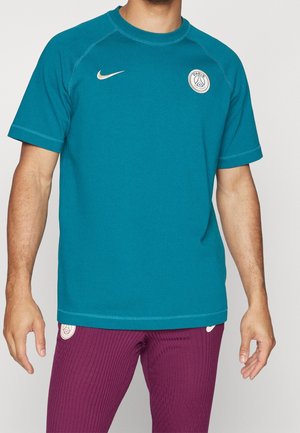 Mężczyzna w turkusowej koszulce Nike Paris Saint-Germain z krótkim rękawem i teksturowanych fioletowych spodniach sportowych na jednolitym tle.