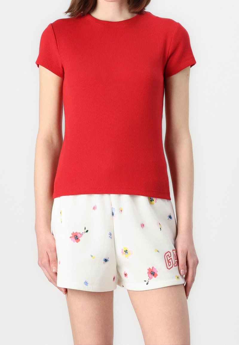 Woman wearing a red short-sleeve top and white floral shorts with "GAP" logo on leg, standing against a white background.