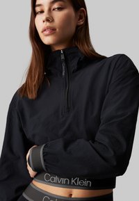 Czarny sweter z wysokim kołnierzem i ćwierć zamkiem, z teksturowaną powierzchnią. Widoczny szary pas Calvin Klein.