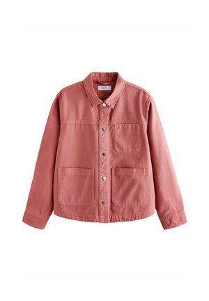 Femme portant une veste rose, un haut blanc, un pantalon large couleur crème et des sandales beige, debout contre un mur en briques et blanc à l'intérieur.